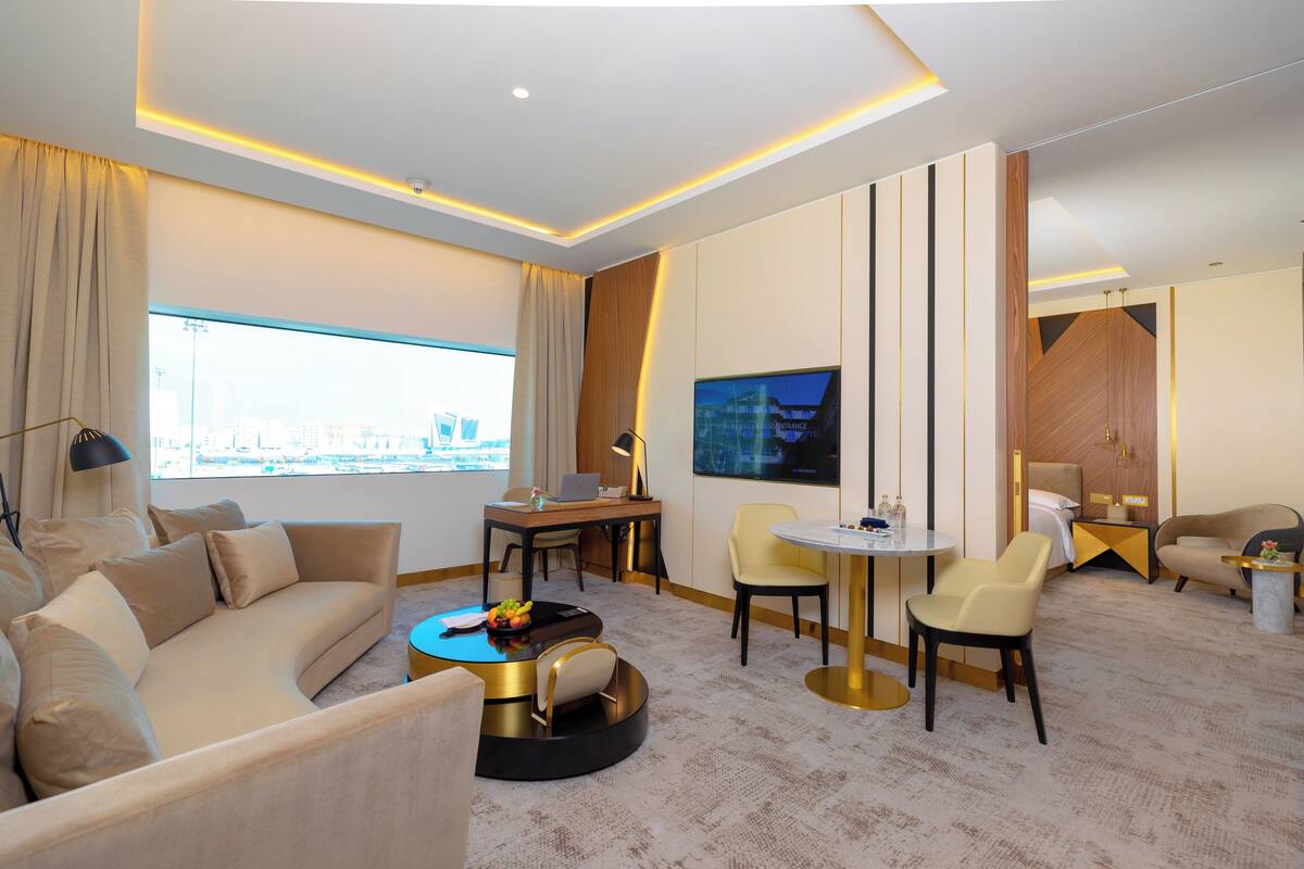 Una habitación de hotel moderna con sofá, mesa de centro redonda, mesa de comedor y sillas. Hay un televisor montado en la pared y un ventanal con vistas a la ciudad.