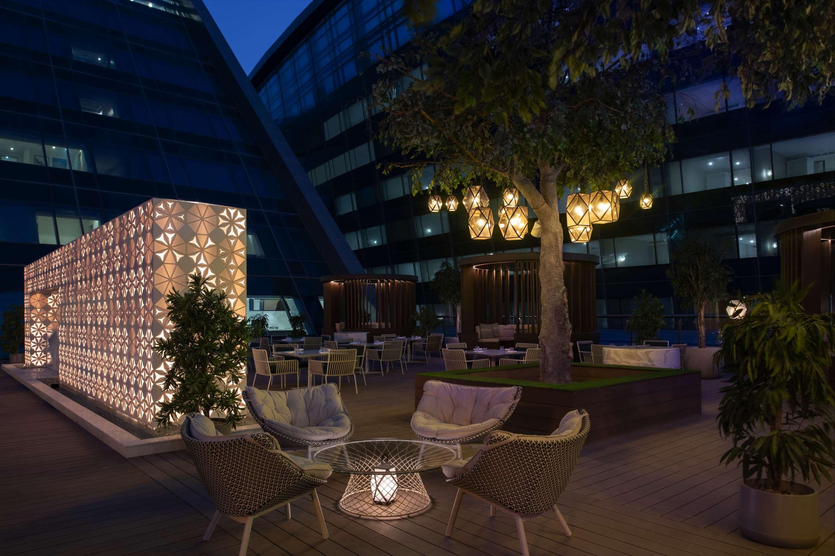 Un área de estar al aire libre con sillas de mimbre y una mesa de vidrio, rodeada por un muro decorativo y árboles.