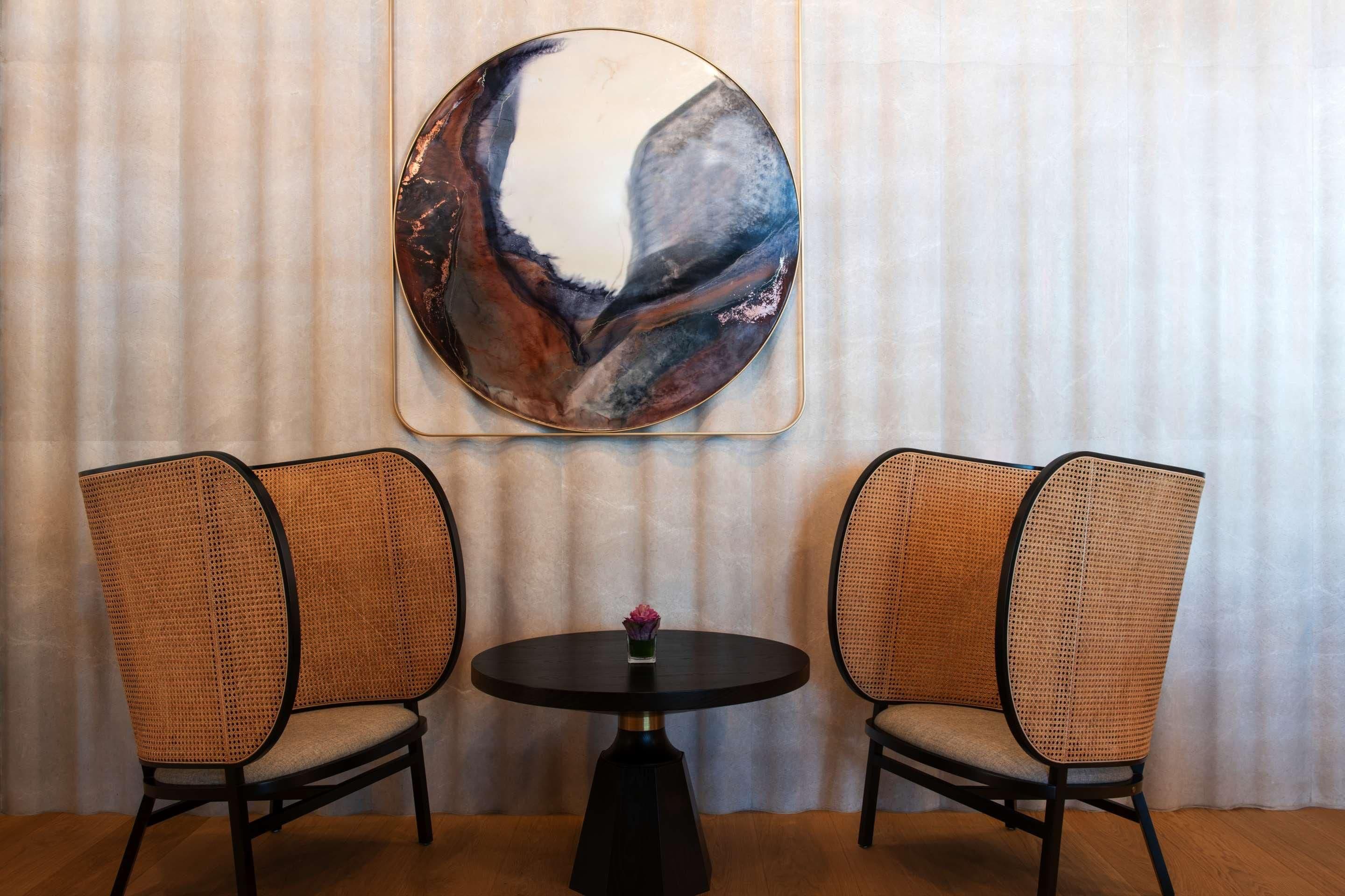 A room with two wicker chairs, a round table with a vase, and a large abstract painting on the wall.