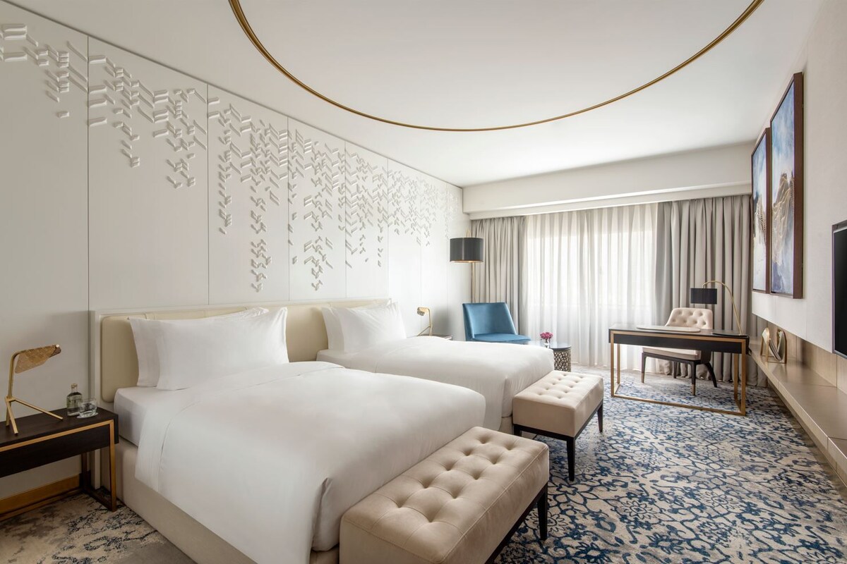 A hotel room with two beds, a desk, a chair, a TV, and a decorative wall.