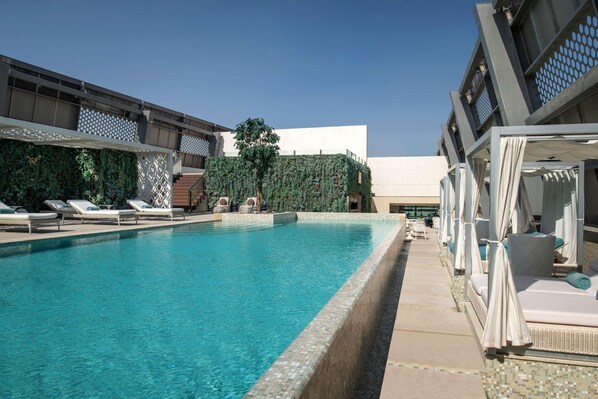 Una piscina con sillones y sombrillas, rodeada por un edificio moderno con fachada geométrica y un muro verde.