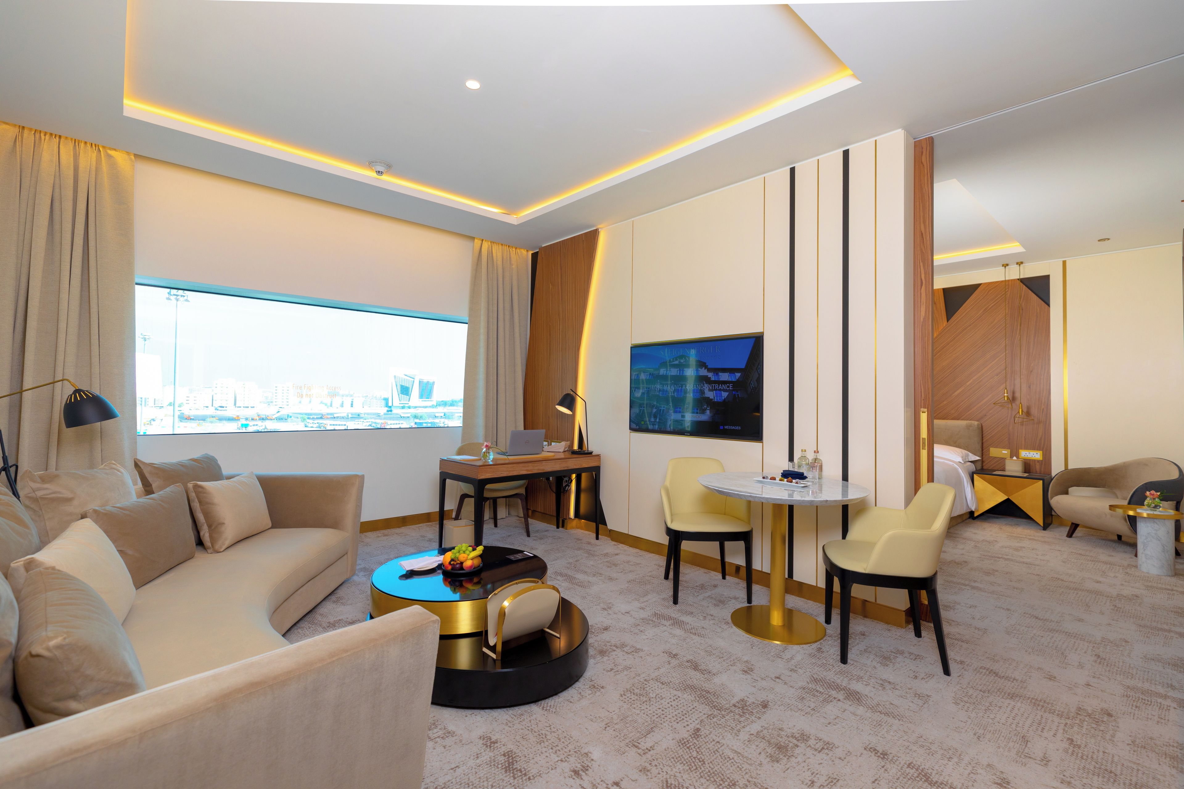 Una habitación de hotel moderna con sofá, mesa de centro redonda, mesa de comedor y sillas. Hay un televisor montado en la pared y un ventanal con vistas a la ciudad.