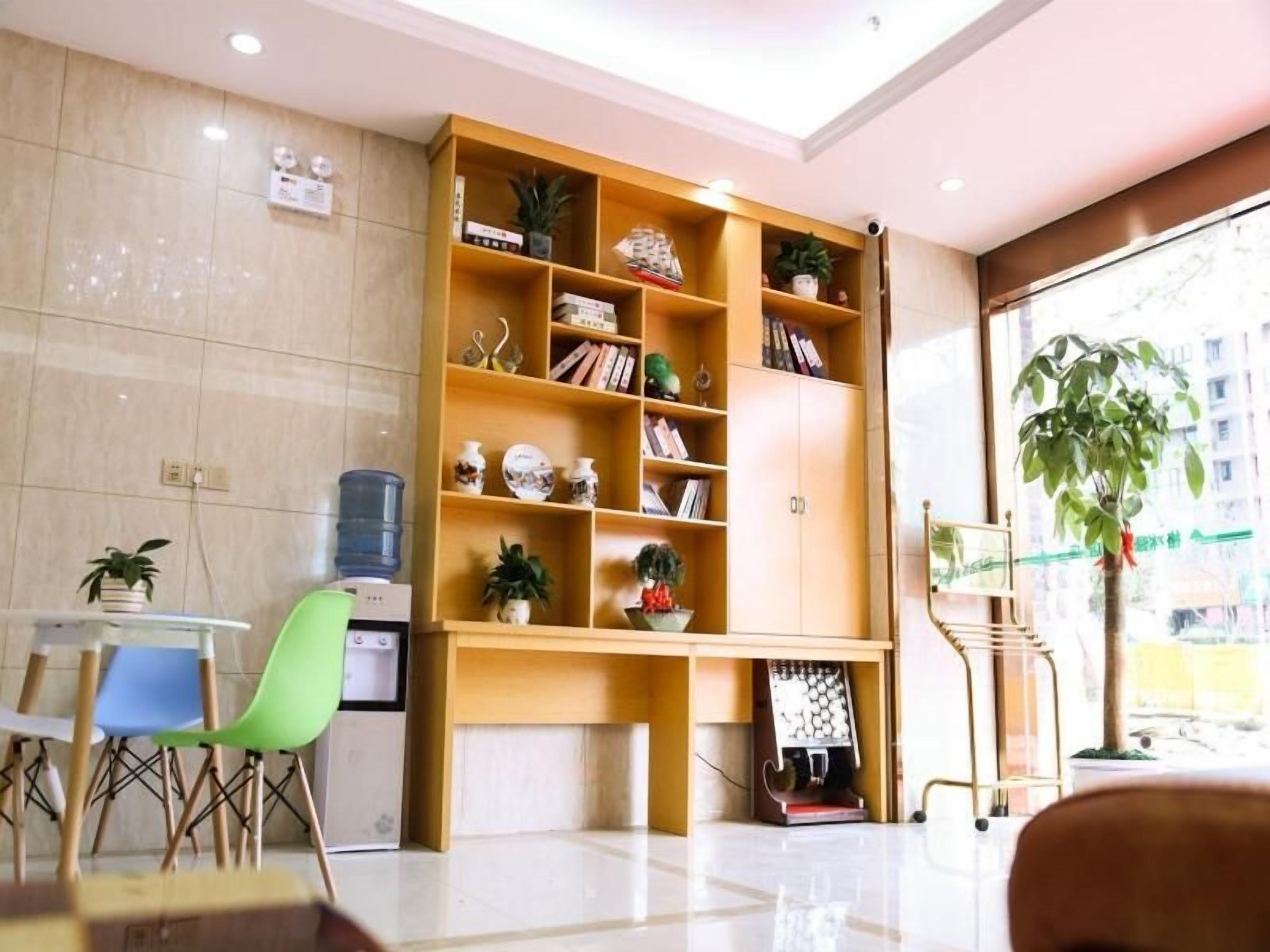 A reception area with a wooden bookshelf, a water dispenser, a green chair, and a large window with a plant.