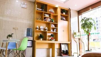 A reception area with a wooden bookshelf, a water dispenser, a green chair, and a large window with a plant.