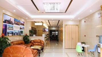 A hotel lobby with a reception desk, seating area with sofas, a dining table with chairs, and a corridor leading to other rooms.