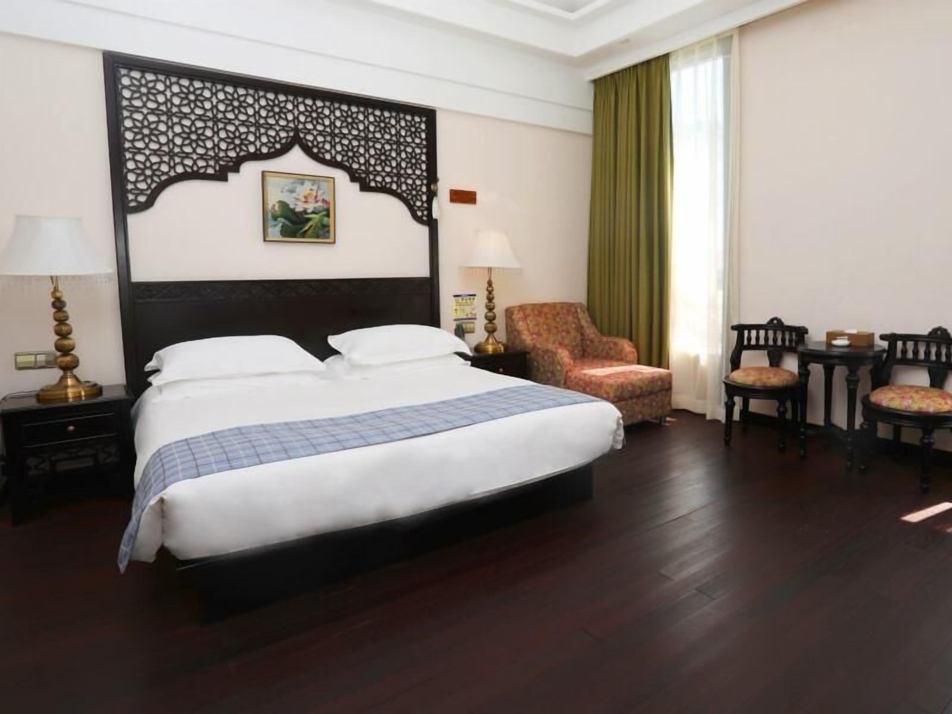 Una habitación de hotel con una cama grande, dos mesitas de noche con lámparas, un sillón floral, dos sillas de madera y un cabecero decorativo.