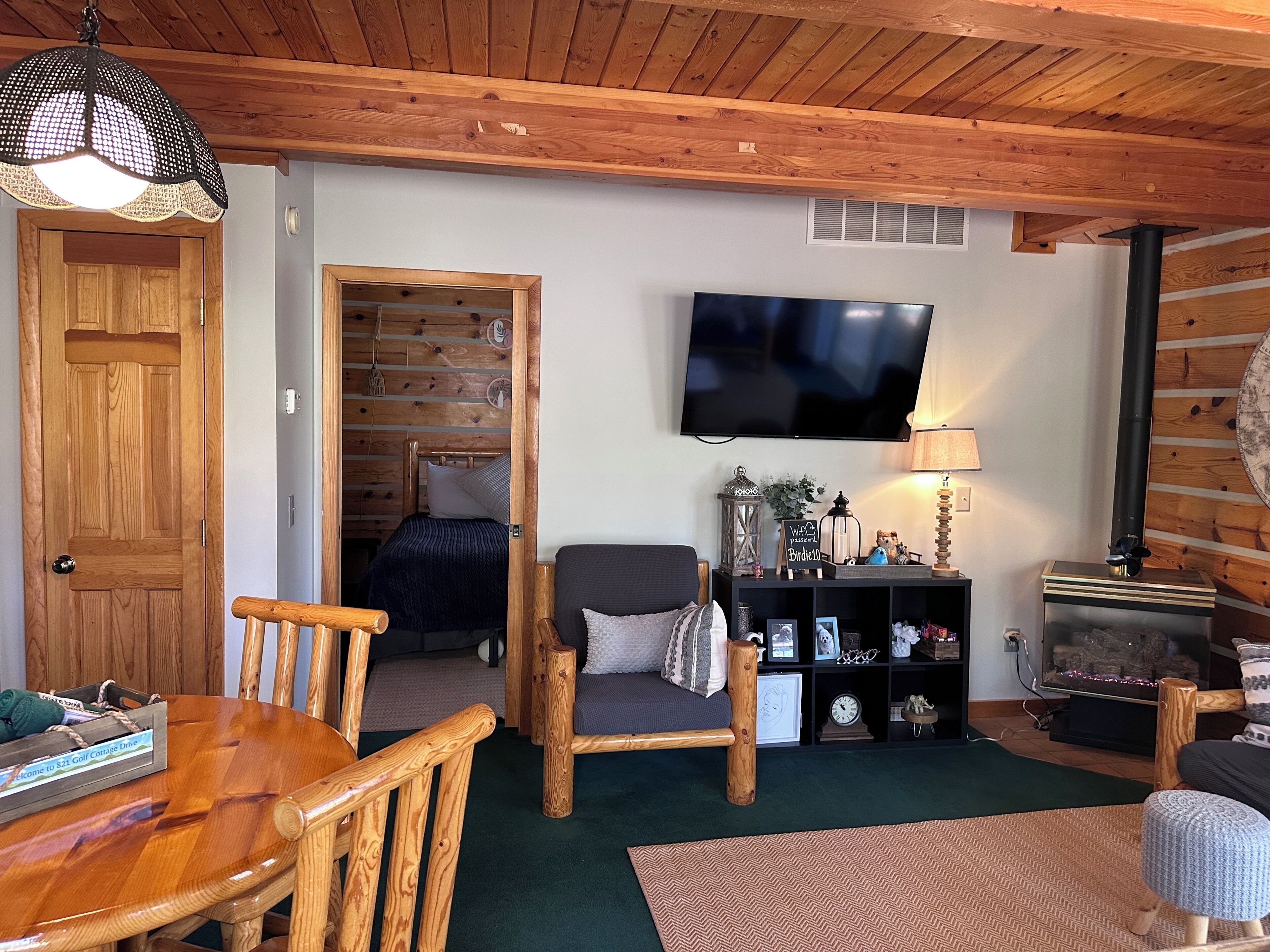Una sala de estar acogedora con techo de madera, un televisor montado en la pared, una chimenea y un comedor con mesa y sillas de madera.