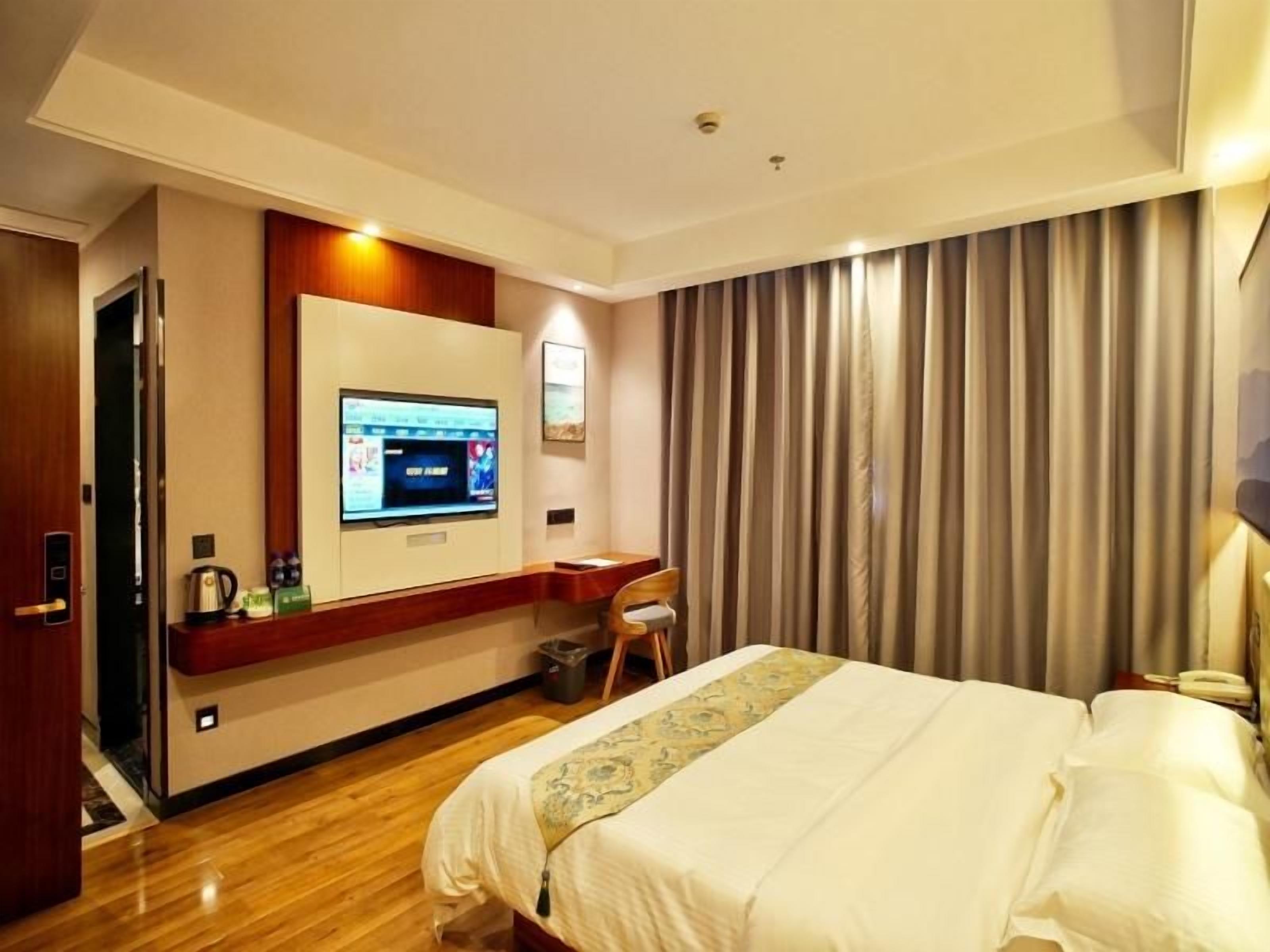 A hotel room with a bed, a television mounted on the wall, a desk with a chair, and a curtain-covered window.