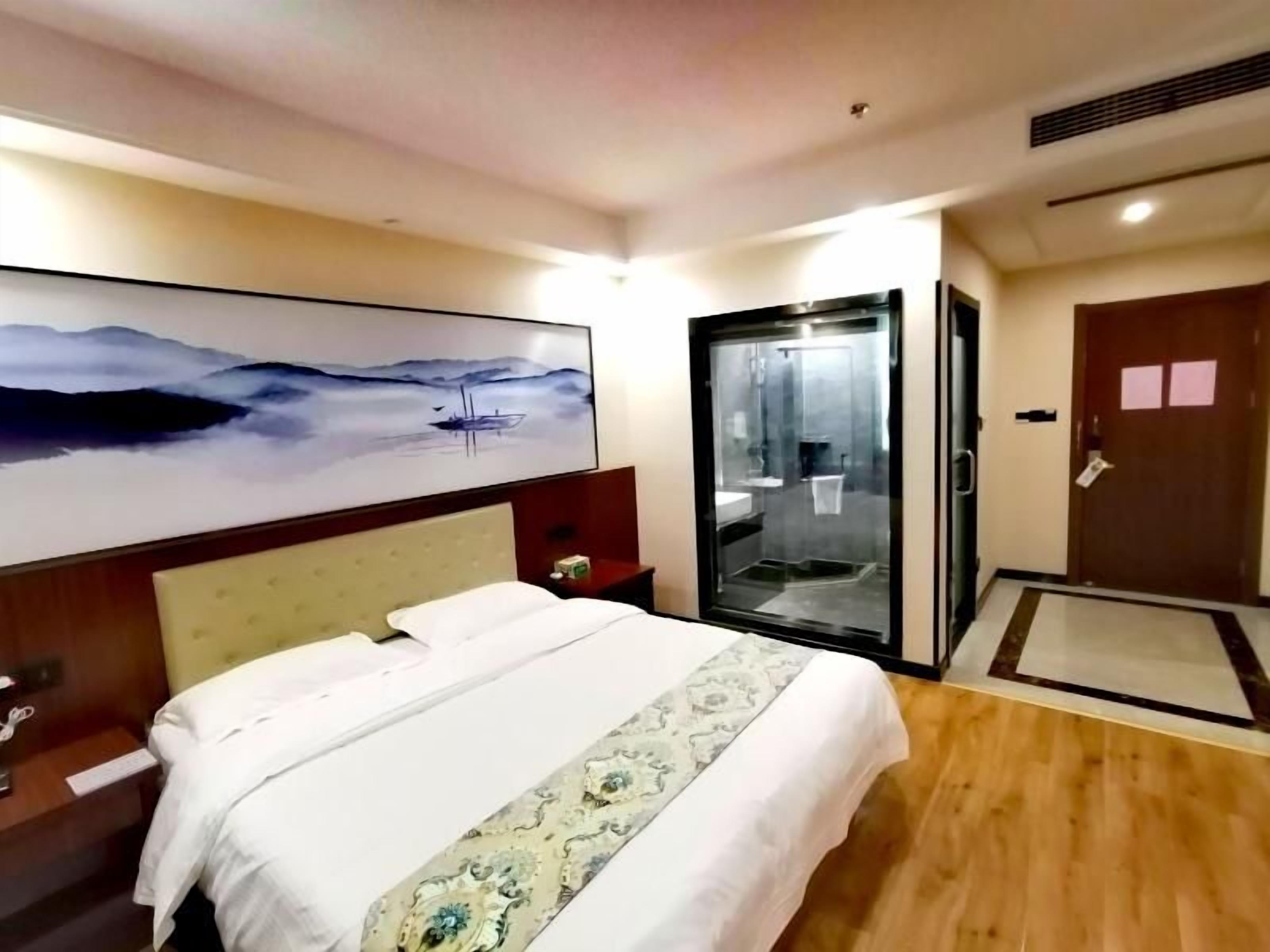 A hotel room with a bed, a painting, a sliding door, and a wooden floor.