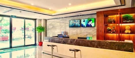 A modern hotel lobby with a reception desk, two bar stools, a large potted plant, and a wall-mounted TV.