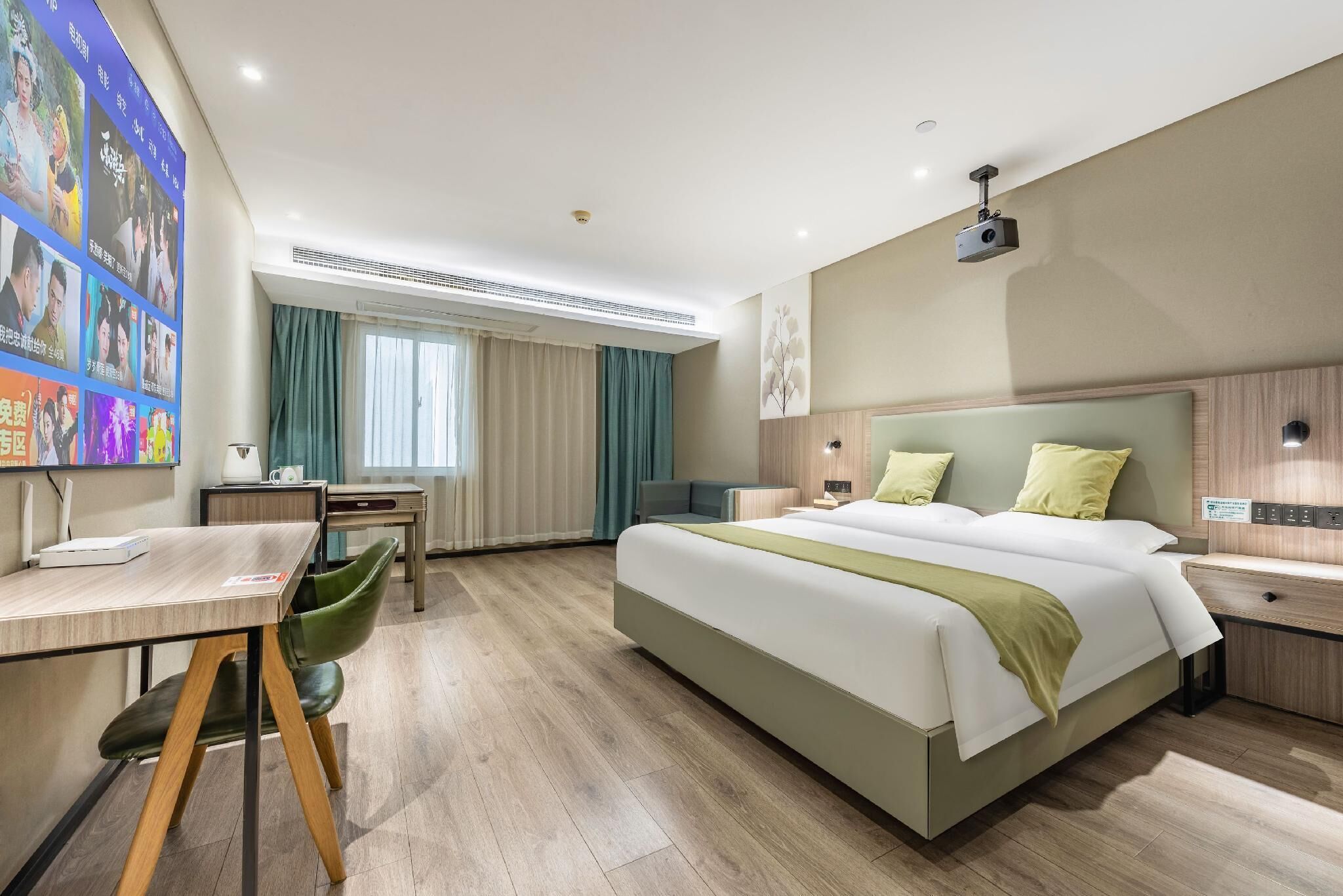 Ein Hotelzimmer mit einem großen Bett, einem Schreibtisch, einem Stuhl, einem kleinen Tisch, einem Projektor und einer Wand mit Fotos.