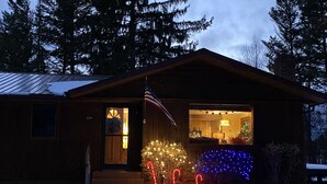 A wooden cabin with a snow-covered roof, a front porch, and a lit Christmas tree. - Swan Mntn View--Entire, Private Basement Suite, Close to GNP, Whitefish & Lake. (Kalispell)