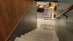 A wooden staircase with a handrail, leading to a lower level. There are framed pictures on the wall, a small table with a chair, and a window with blinds. - Swan Mntn View--Entire, Private Basement Suite, Close to GNP, Whitefish & Lake. (Kalispell)