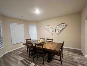 Comedor con mesa y sillas de madera, dos adornos en la pared y una ventana con persianas.