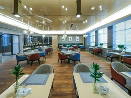 A spacious dining area with multiple tables and chairs, large windows with blinds, and ceiling lights.