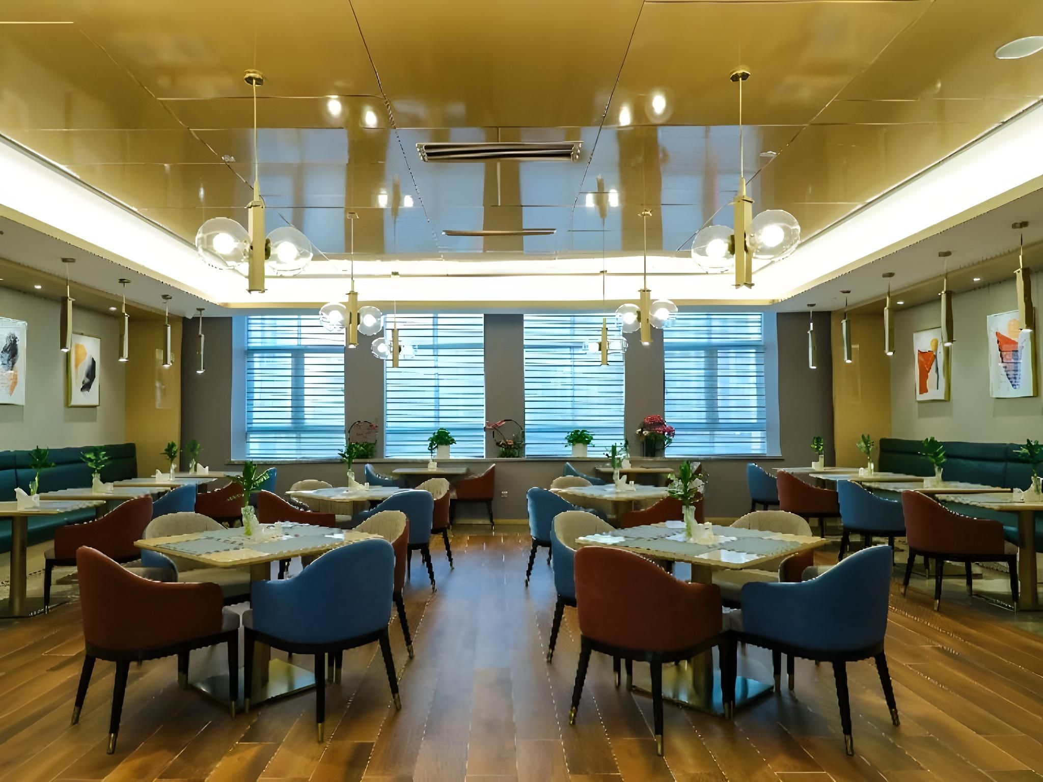 A spacious restaurant with multiple tables and chairs, large windows with blinds, and hanging pendant lights.