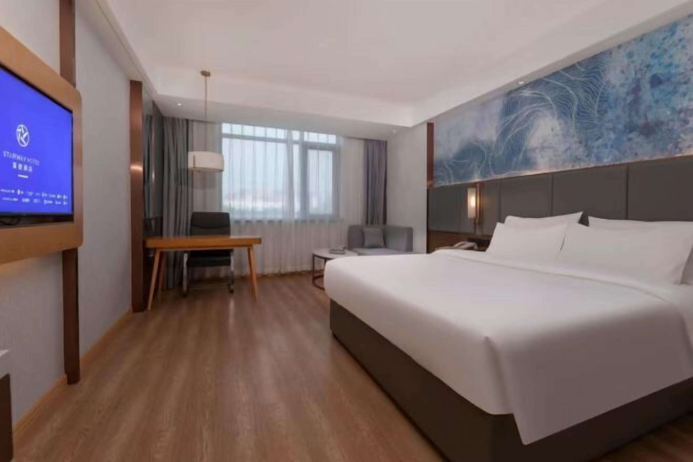 A hotel room with a large bed, a desk, a chair, a TV, and a window with curtains.
