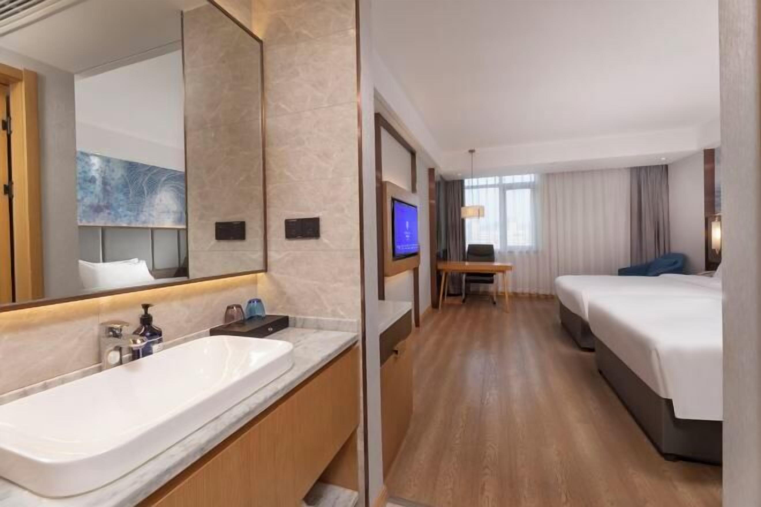 A hotel room with a large bathroom sink, a bed, a desk with a chair, and a television.