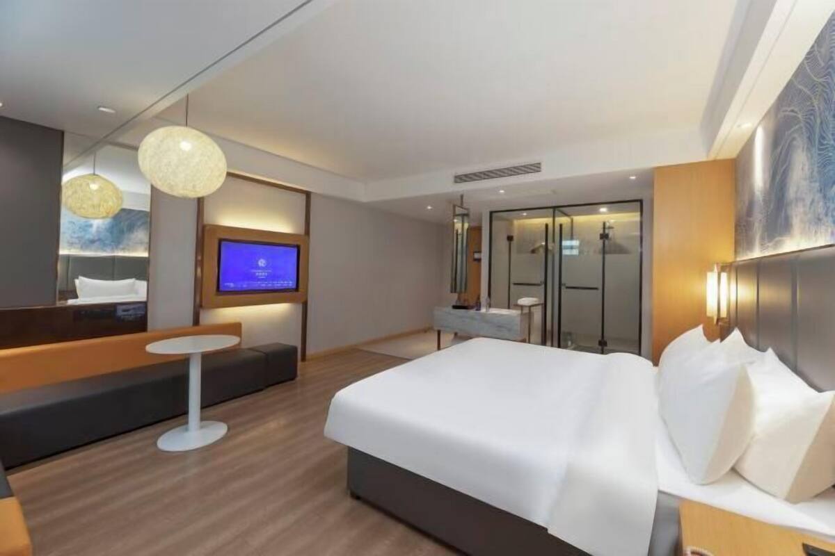 A modern hotel room with a large bed, a small round table, a sofa, a TV, and a glass-enclosed shower area.