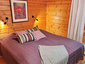 Quarto com paredes de madeira, cama com colcha roxa, travesseiros listrados, um cobertor e um quadro na parede.