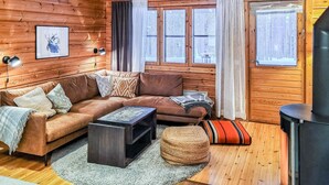 A cozy log cabin interior with a brown leather sofa, a small coffee table, and a woven ottoman. There are two windows with curtains, a clock on the wall, and a framed picture above the sofa. - Purnukka by Interhome (Kuusamo)