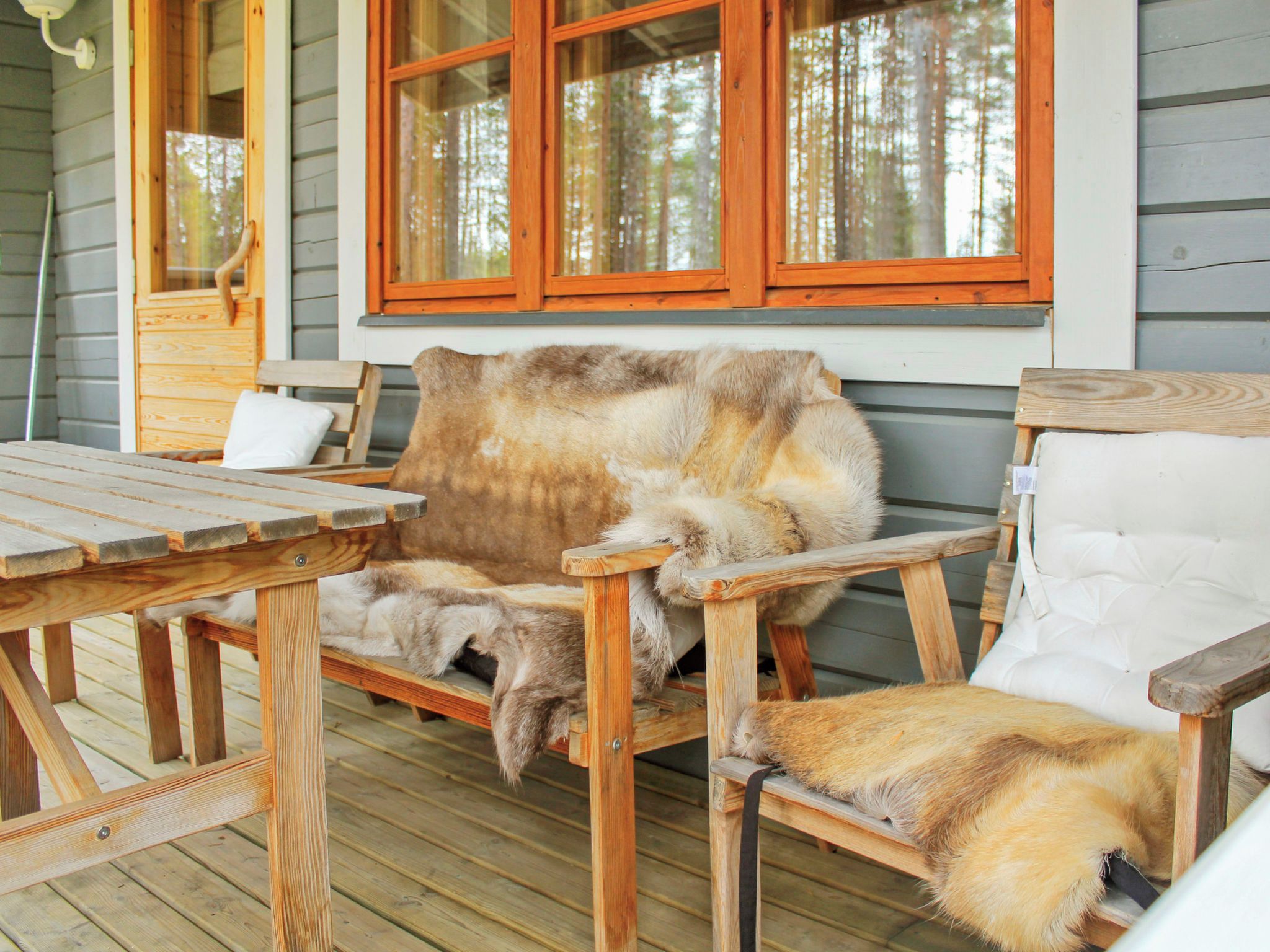 Puisesta laudasta rakennettu kuisti, jossa on penkki ja pöytä, ikkuna, josta avautuu näkymä puihin, ja valkoinen, pehmustettu tuoli.