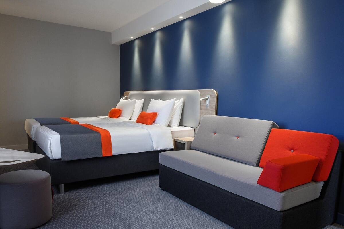 A modern hotel room with two beds, a grey sofa, and a small table.