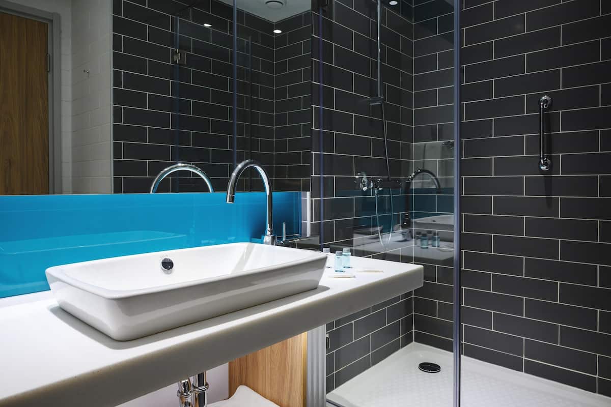 A modern bathroom with a large white sink, a glass shower enclosure, and dark tiled walls.