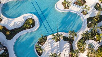 Vue aérienne d’un complexe hôtelier doté d’une grande piscine centrale, de plusieurs piscines plus petites et d’espaces de détente aménagés tout autour, avec des palmiers.