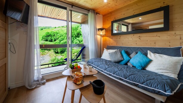 Een knus interieur met houten plafond, groot raam met uitzicht op groen, bank met kussens, tafel met een mand brood, en een vloerlamp.