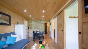 Een knus vertrek met een houten plafond, een bank met blauwe kussens, een eettafel met stoelen en een kleine keukenhoek.