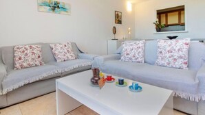 Una sala de estar con dos sofás grises, cada uno con cojines florales, una mesa de centro blanca con tazas de colores, y un cuadro en la pared.