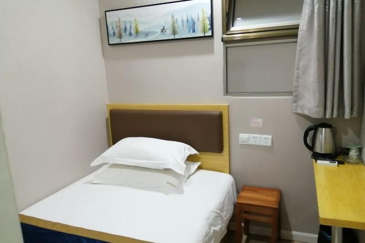 Quarto de hotel com uma cama, um banquinho de madeira, uma mesa com chaleira e um quadro na parede.