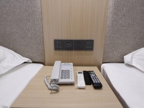 Habitació d'hotel amb una taula de nit de fusta, un telèfon, un comandament a distància i una presa elèctrica.