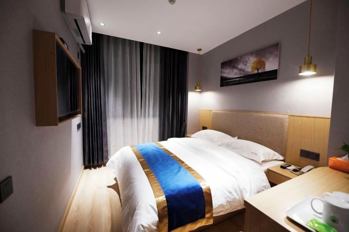A hotel room with a bed, a bedside table, a nightstand, a wall-mounted TV, and a window with curtains.