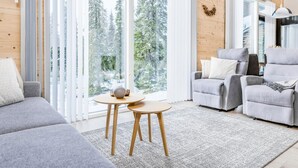 Een moderne woonkamer met een grijze bank, een grijze fauteuil en een kleine ronde tafel. Grote ramen met witte gordijnen bieden uitzicht op een bebost gebied.