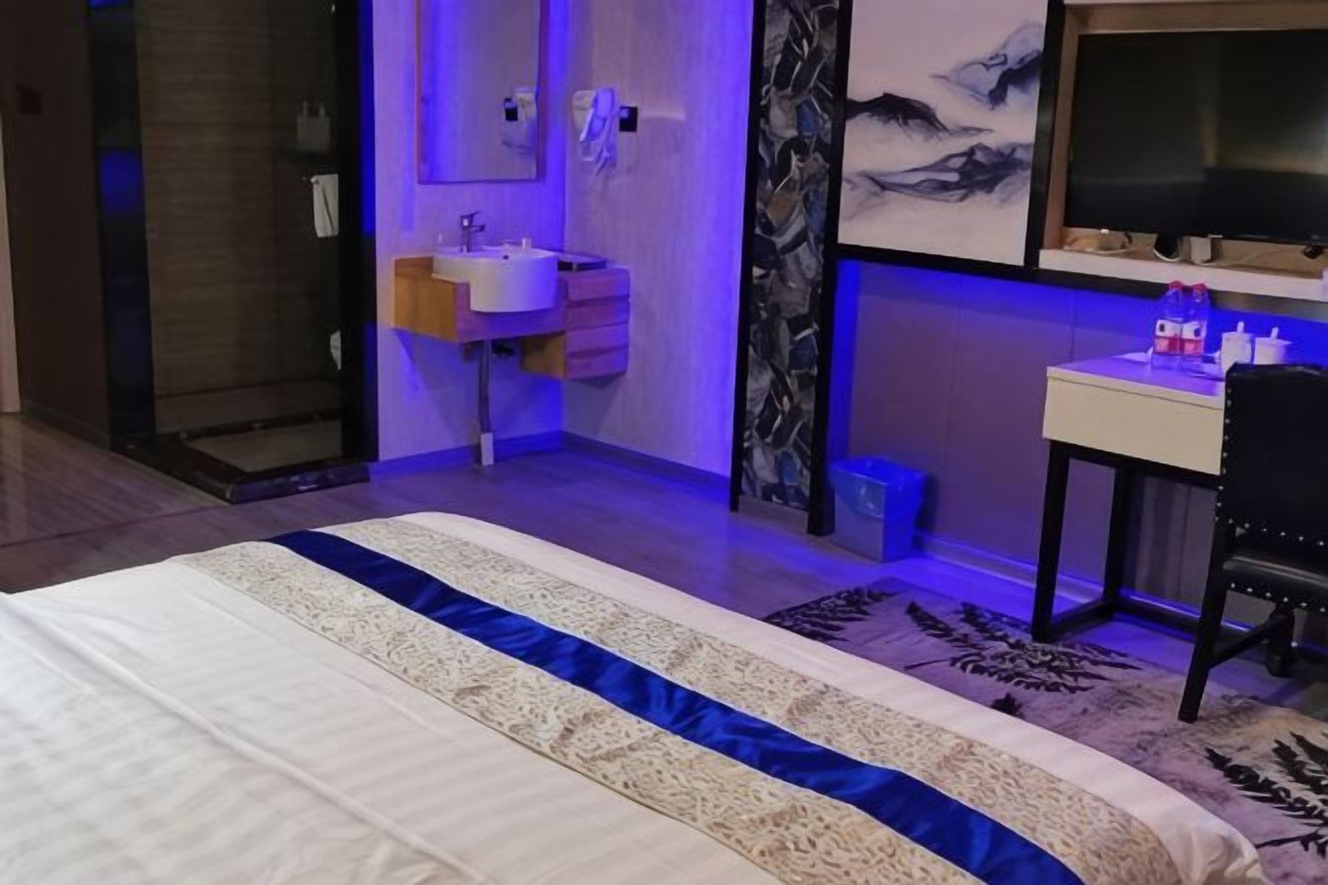 A bedroom with a bed, a bedside table, a nightstand, a window, and a bathroom with a sink and mirror.