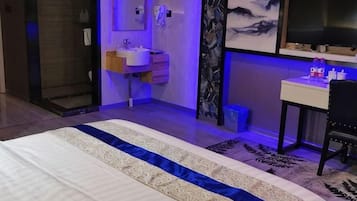 A bedroom with a bed, a bedside table, a nightstand, a window, and a bathroom with a sink and mirror.