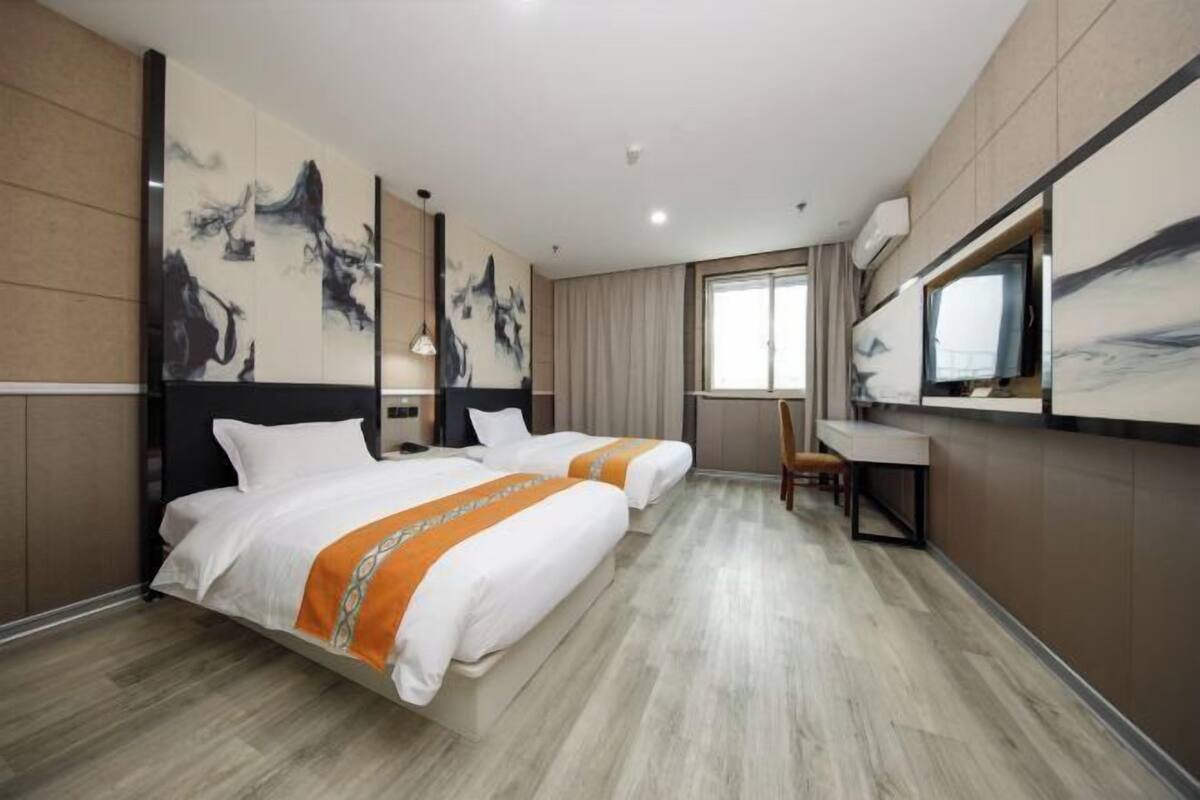 A hotel room with two beds, a desk, a chair, a large window, and artwork on the walls.