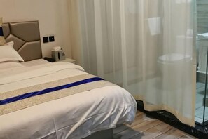 Habitación de hotel con cama, mesita de noche, lámpara y vistas a través de cortinas translúcidas.