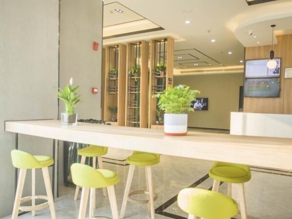Uma área de recepção com balcão, banquetas verdes, plantas em vasos e uma tela de televisão.