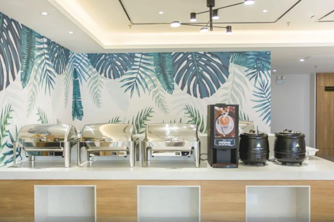 Un mostrador de buffet con un mural de hojas tropicales, una máquina de café y dos hervidores negros.