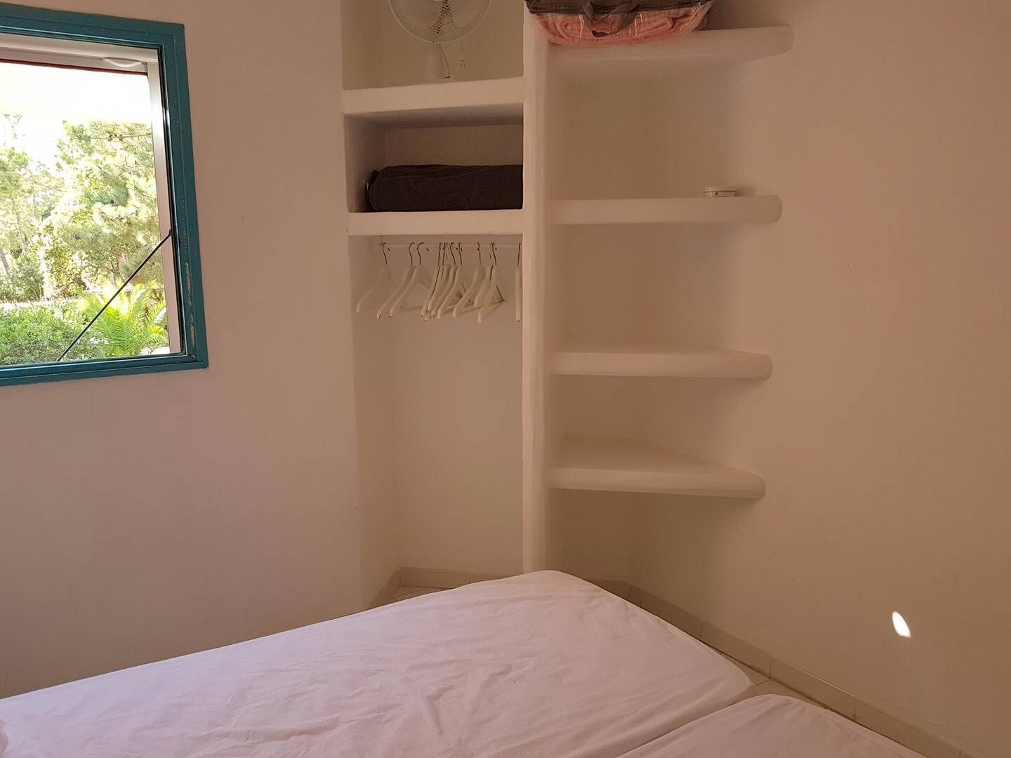 A bedroom with a bed, a window with a view of trees, and a white wall with a shelf and a fan.