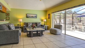 A living room with a sofa, armchair, coffee table, and a glass door leading to an outdoor patio. - Disney Launchpad for Families! Private Pool (Kissimmee)