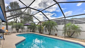 A covered swimming pool with a clear blue water, surrounded by lounge chairs and umbrellas, and a view of palm trees and a house in the background. - Disney Launchpad for Families! Private Pool (Kissimmee)