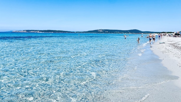 En strand med klart vand, folk der går langs kysten og fjerne bakker.