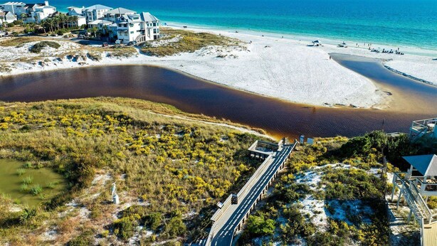 Vue aérienne d’une zone côtière comprenant une plage, une étendue d’eau et une route menant à un ensemble de bâtiments.