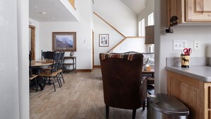 Zona pranzo con tavolo e sedie in legno, una scala che conduce a un livello superiore e una cucina con pensili e un piano di lavoro in legno.