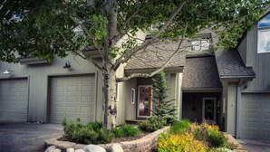 A residential house with two garage doors, a front porch with a red door, and a landscaped garden with rocks and shrubs. - 3BR Pet Friendly - Ski Hill Rd (Driggs)
