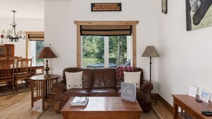 A cozy living room with a leather sofa, wooden coffee table, and a dining area with a wooden table and chairs. A large window offers a view of the outdoors. - 3BR Pet Friendly - Ski Hill Rd (Driggs)